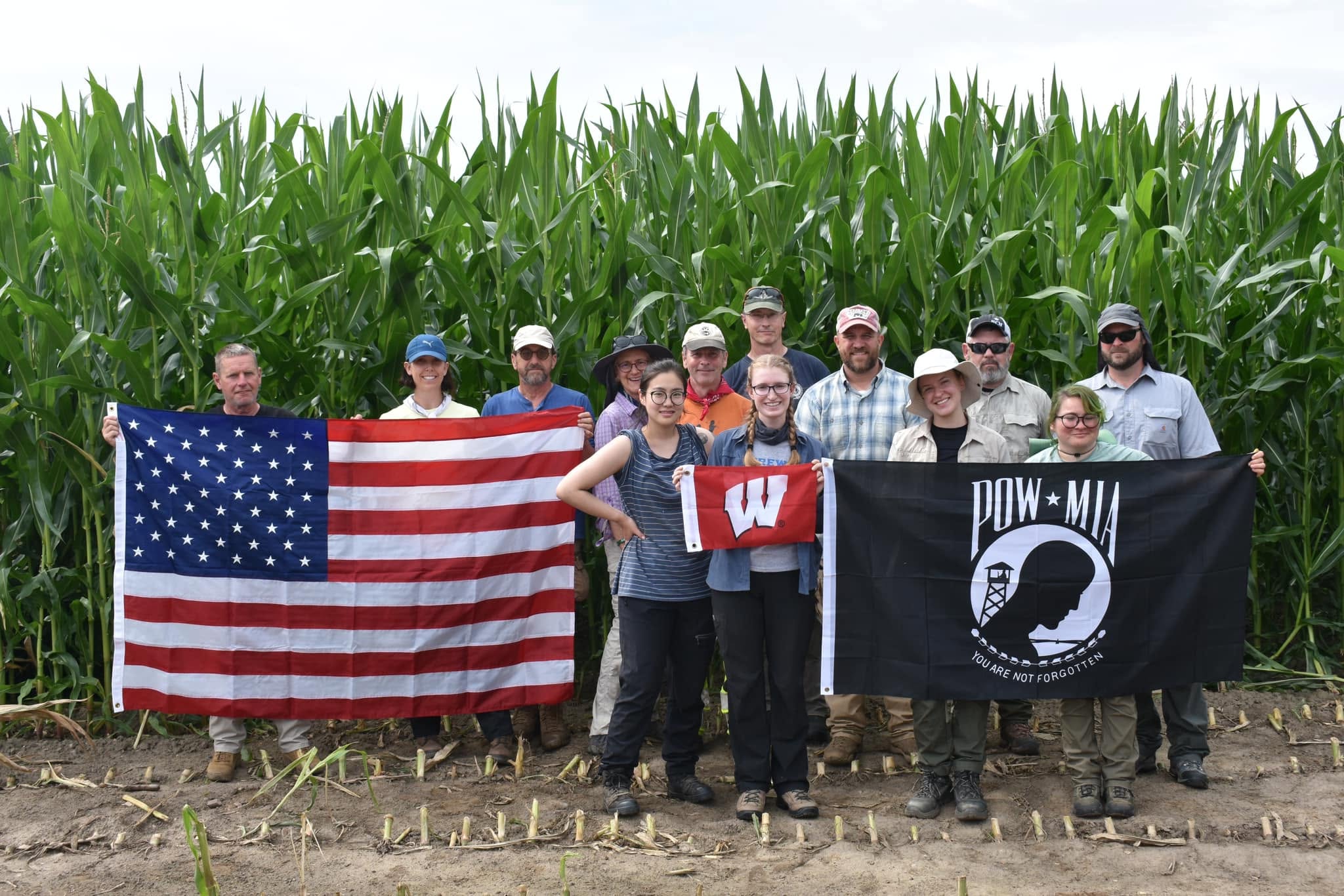 UW-MIA recovery team at a field excavation site, holding the American flag, the University of Wisconsin flag, and the POW/MIA flag